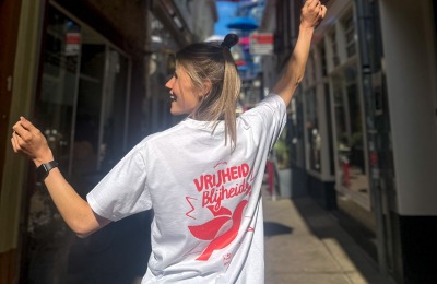Vrouw met blond haar draagt een wit T-shirt met opdruk 'Vrijheid Blijheid' in een smalle straat.