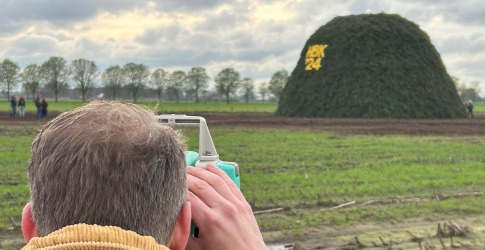 Een man in een corduroy jas kijkt door een meetinstrument naar een grote groene stapel met 'BX 24'.