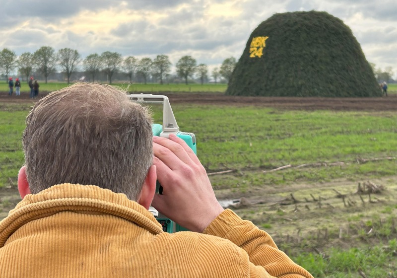 Een man in een corduroy jas kijkt door een meetinstrument naar een grote groene stapel met 'BX 24'.