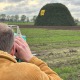 Een man in een corduroy jas kijkt door een meetinstrument naar een grote groene stapel met 'BX 24'.