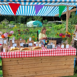 Kraam met rood-wit geruit tafelkleed op een markt, bloemen, producten en twee mensen op de achtergrond.