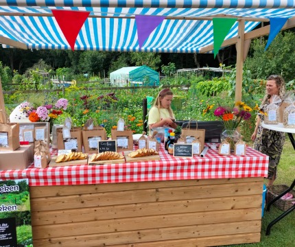 Kraam met rood-wit geruit tafelkleed op een markt, bloemen, producten en twee mensen op de achtergrond.