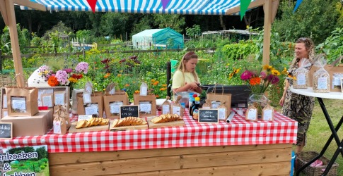 Kraam met rood-wit geruit tafelkleed op een markt, bloemen, producten en twee mensen op de achtergrond.