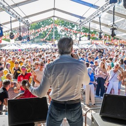 Zanger op een podium met microfoon kijkt uit op een groot publiek op een zonnig evenement in een tent.