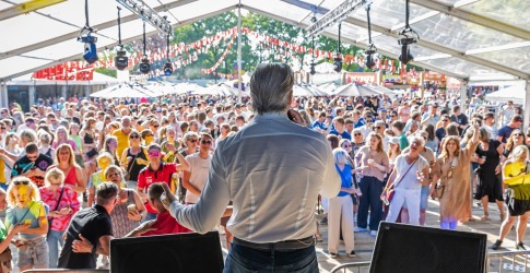 Zanger op een podium met microfoon kijkt uit op een groot publiek op een zonnig evenement in een tent.