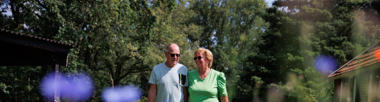 Twee oudere volwassenen wandelen samen door een groene tuin met paarse bloemen op een zonnige dag.