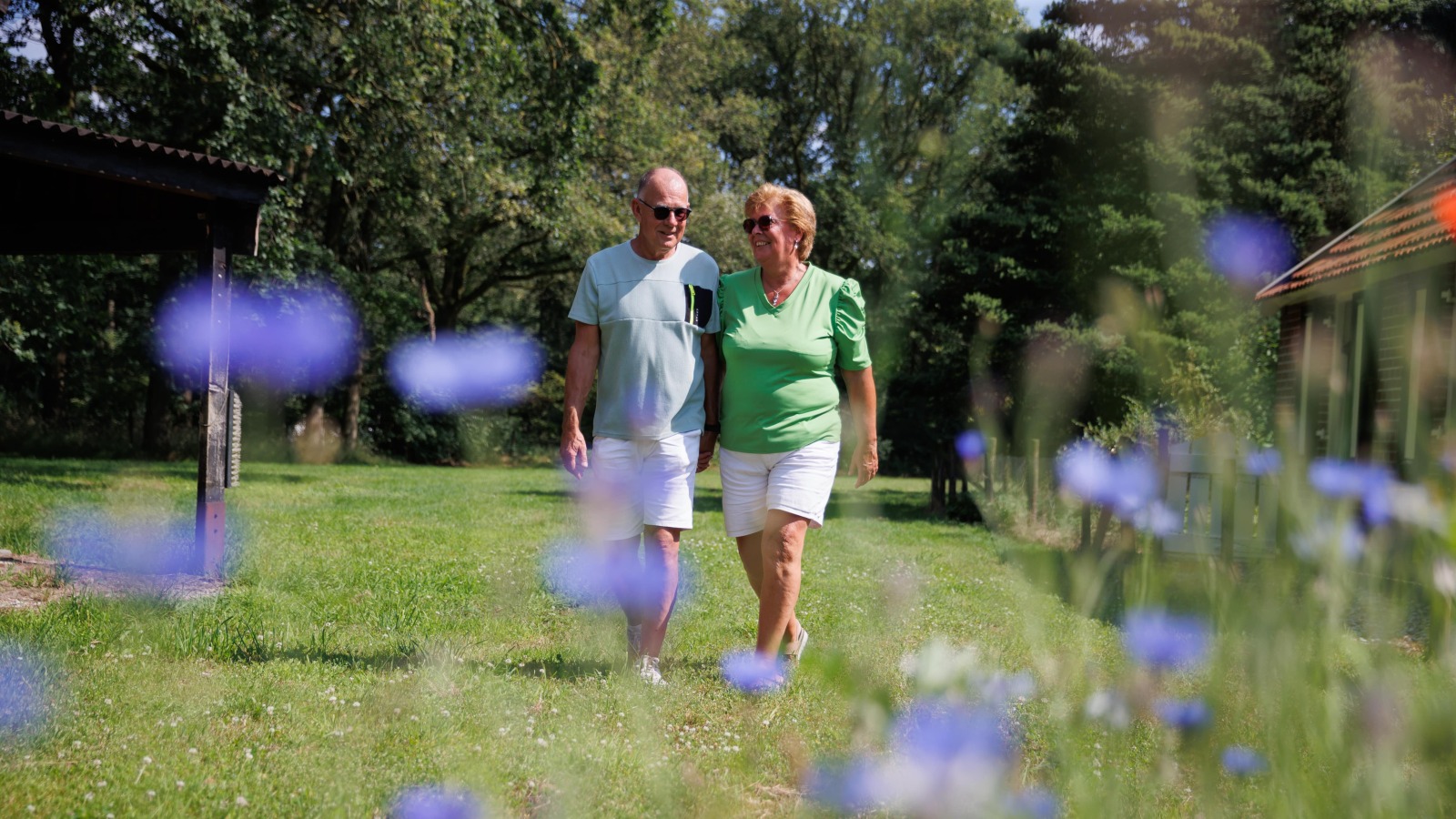 Twee oudere volwassenen wandelen samen door een groene tuin met paarse bloemen op een zonnige dag.