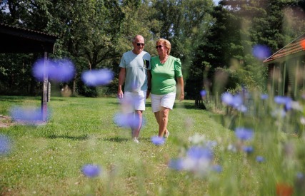 Twee oudere volwassenen wandelen samen door een groene tuin met paarse bloemen op een zonnige dag.