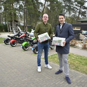 Twee mannen staan buiten met folders, elektrische scooters op de achtergrond, recreatiegebied sfeer.