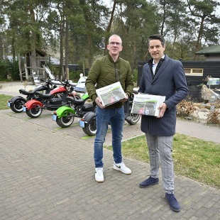 Twee mannen staan buiten met folders, elektrische scooters op de achtergrond, recreatiegebied sfeer.