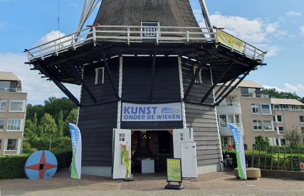 Een traditionele Nederlandse windmolen met een tentoonstelling genaamd 'Kunst onder de Wieken' op een zonnige dag.