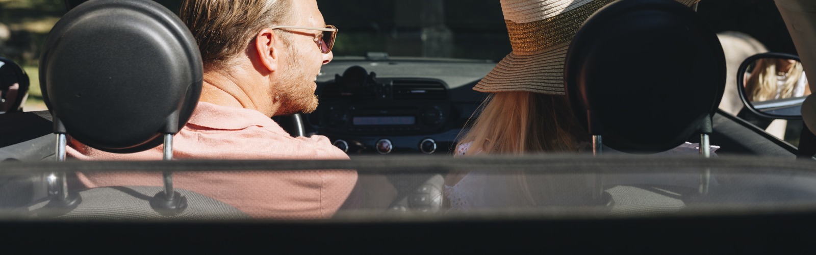 Twee mensen zitten in een cabriolet, de man met zonnebril en de vrouw met een grote hoed op een zonnige dag.