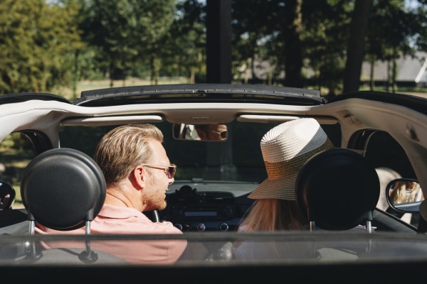 Twee mensen zitten in een cabriolet, de man met zonnebril en de vrouw met een grote hoed op een zonnige dag.