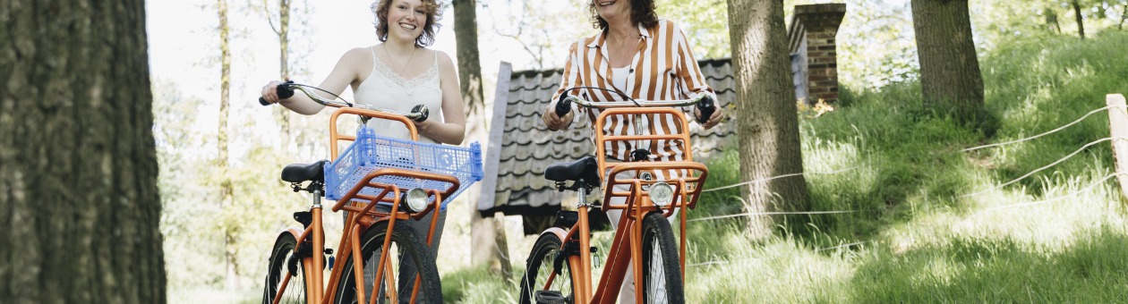 Twee vrouwen lopen met oranje fietsen door een groen bosrijk gebied op een zonnige dag in de natuur.