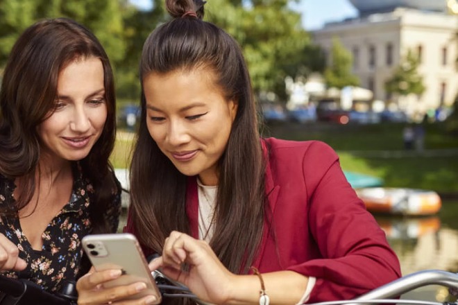 Twee vrouwen op een zonnige dag bij het water kijken samen op een smartphone, naast fietsen.