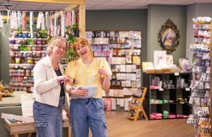 Twee lachende vrouwen staan samen in een kleurrijke hobbywinkel met knutselspullen op de achtergrond.