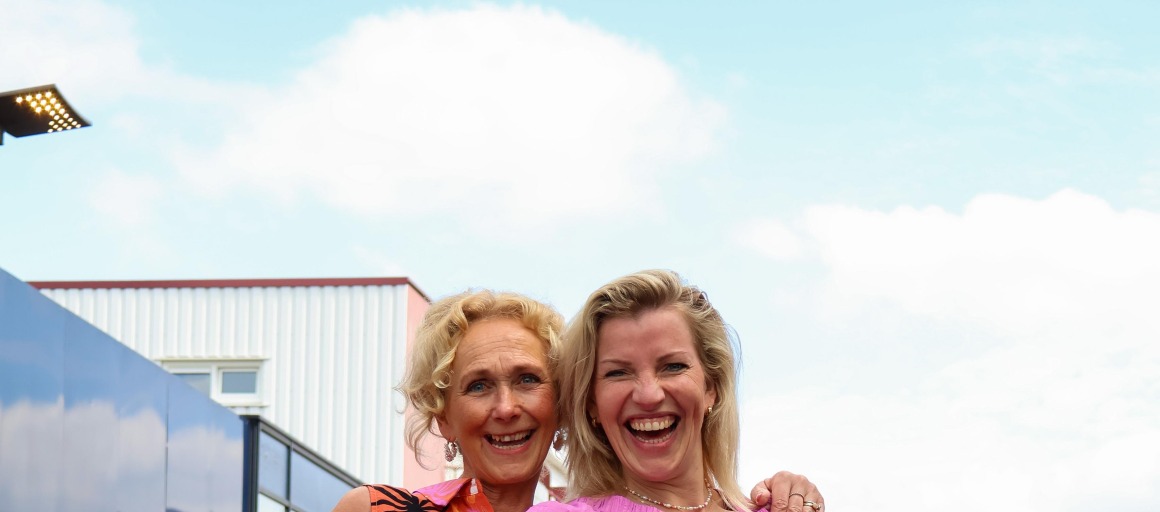 Twee vrouwen lachen en poseren op een catwalk, dragen kleurrijke zomerkleding tijdens een modeshow buiten.