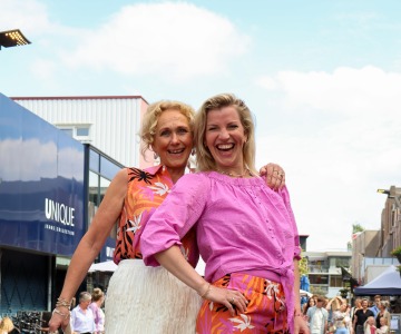 Twee vrouwen lachen en poseren op een catwalk, dragen kleurrijke zomerkleding tijdens een modeshow buiten.