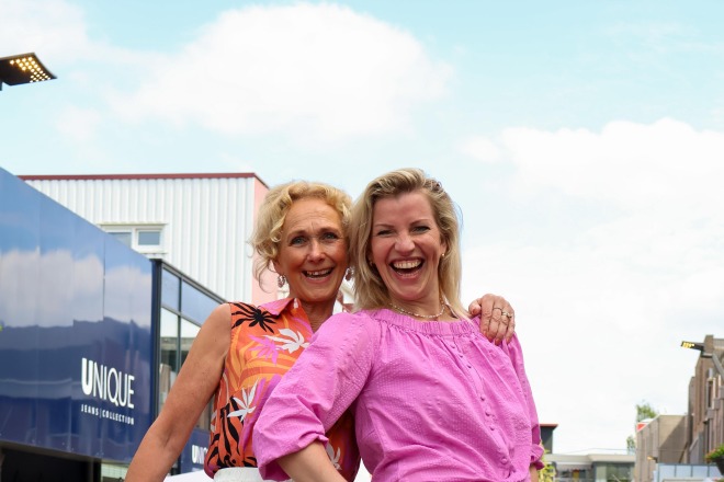Twee vrouwen lachen en poseren op een catwalk, dragen kleurrijke zomerkleding tijdens een modeshow buiten.
