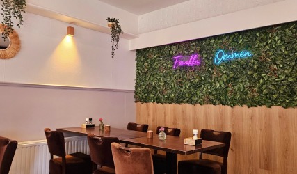 Gezellige caféruimte met bruine stoelen, houten tafels, groene plantenmuur en neonverlichting.
