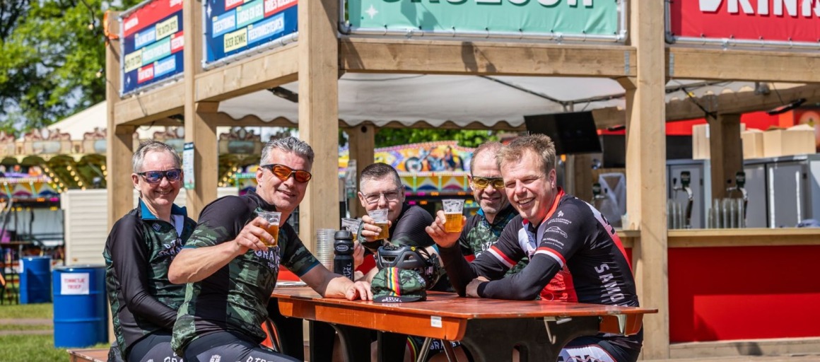 Vijf wielrenners in wielerkleding zitten aan een picknicktafel en genieten van een biertje bij een Grolsch-bar.