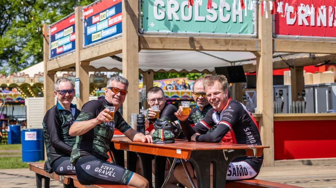 Vijf wielrenners in wielerkleding zitten aan een picknicktafel en genieten van een biertje bij een Grolsch-bar.