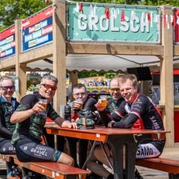 Vijf wielrenners in wielerkleding zitten aan een picknicktafel en genieten van een biertje bij een Grolsch-bar.