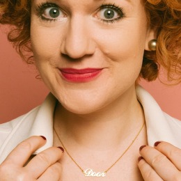 Vrouw met krullend rood haar, opvallende oorbellen en een gouden ketting met de naam 'Door', lachend.