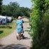Een vrouw loopt op een zandpad met een kinderwagen, omgeven door groen en kampeerterreinen.