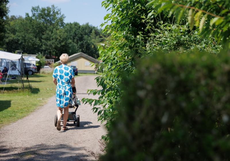 Een vrouw loopt op een zandpad met een kinderwagen, omgeven door groen en kampeerterreinen.