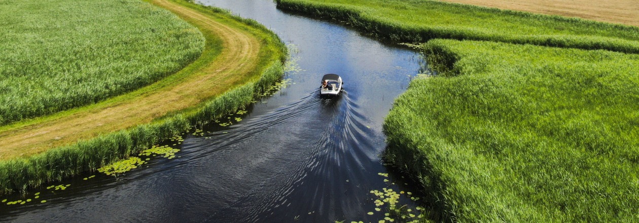Een boot vaart door een kronkelende rivier omringd door groene velden en een blauwe lucht met wolken.