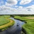 Een boot vaart door een kronkelende rivier omringd door groene velden en een blauwe lucht met wolken.
