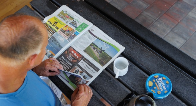 Een man leest een krant op een picknicktafel met een beker, koekjestrommel en koffiekan naast zich.
