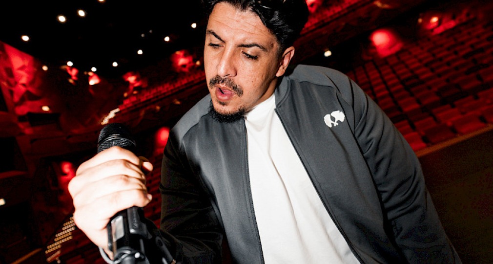 Man houdt een microfoon vast op een podium in een rood verlichte theaterzaal met lege stoelen.