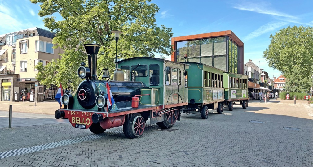Kleurrijke toeristentrein in klassieke stoomlocomotiefstijl op een plein met moderne gebouwen en bomen.