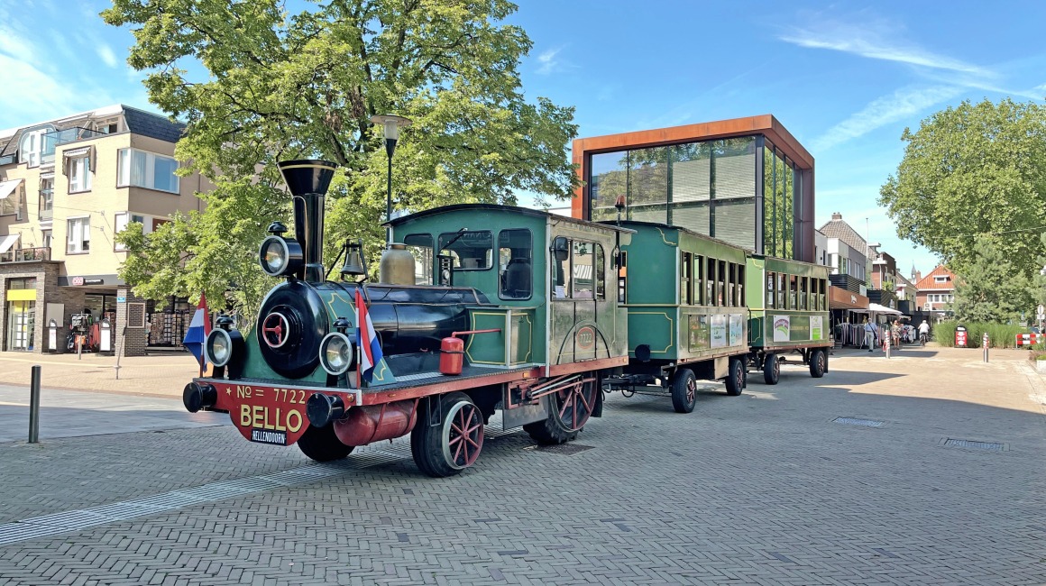Kleurrijke toeristentrein in klassieke stoomlocomotiefstijl op een plein met moderne gebouwen en bomen.