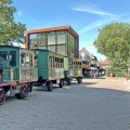 Kleurrijke toeristentrein in klassieke stoomlocomotiefstijl op een plein met moderne gebouwen en bomen.