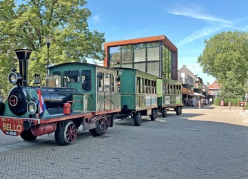 Kleurrijke toeristentrein in klassieke stoomlocomotiefstijl op een plein met moderne gebouwen en bomen.