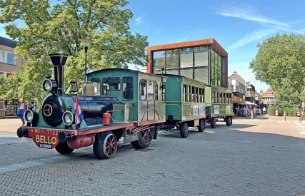 Kleurrijke toeristentrein in klassieke stoomlocomotiefstijl op een plein met moderne gebouwen en bomen.