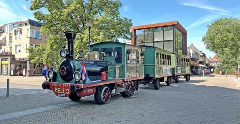 Kleurrijke toeristentrein in klassieke stoomlocomotiefstijl op een plein met moderne gebouwen en bomen.