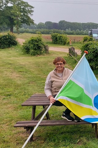 Vrouw zit op een picknicktafel met een kleurrijke vlag op een landelijke camping met campers op de achtergrond.