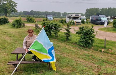 Vrouw zit op een picknicktafel met een kleurrijke vlag op een landelijke camping met campers op de achtergrond.