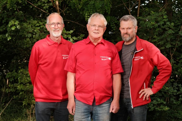Drie oudere mannen poseren samen buiten voor groene struiken, gekleed in rode shirts of jas.