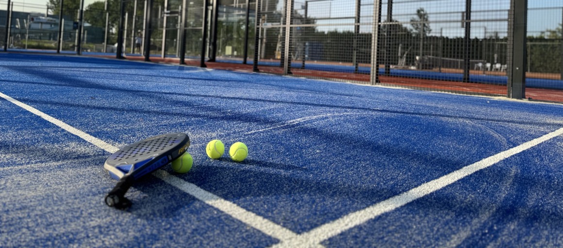 Padelbaan met blauw kunstgras, drie gele ballen en een racket op de grond op een zonnige dag.