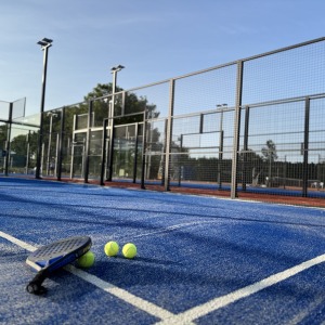 Padelbaan met blauw kunstgras, drie gele ballen en een racket op de grond op een zonnige dag.