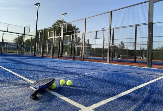 Padelbaan met blauw kunstgras, drie gele ballen en een racket op de grond op een zonnige dag.