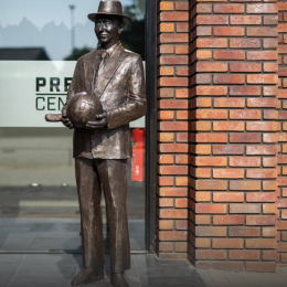 Standbeeld van een man in pak en hoed die een voetbal vasthoudt, geplaatst bij een bakstenen muur.