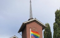 Een bakstenen kerk met een regenboogvlag gehesen aan een vlaggenmast, omgeven door groen gras.
