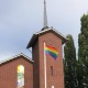 Een bakstenen kerk met een regenboogvlag gehesen aan een vlaggenmast, omgeven door groen gras.
