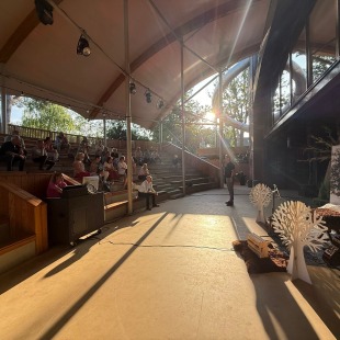 Een zonovergoten amfitheater met houten zitplaatsen, publiek en een spreker op het podium in de natuur.